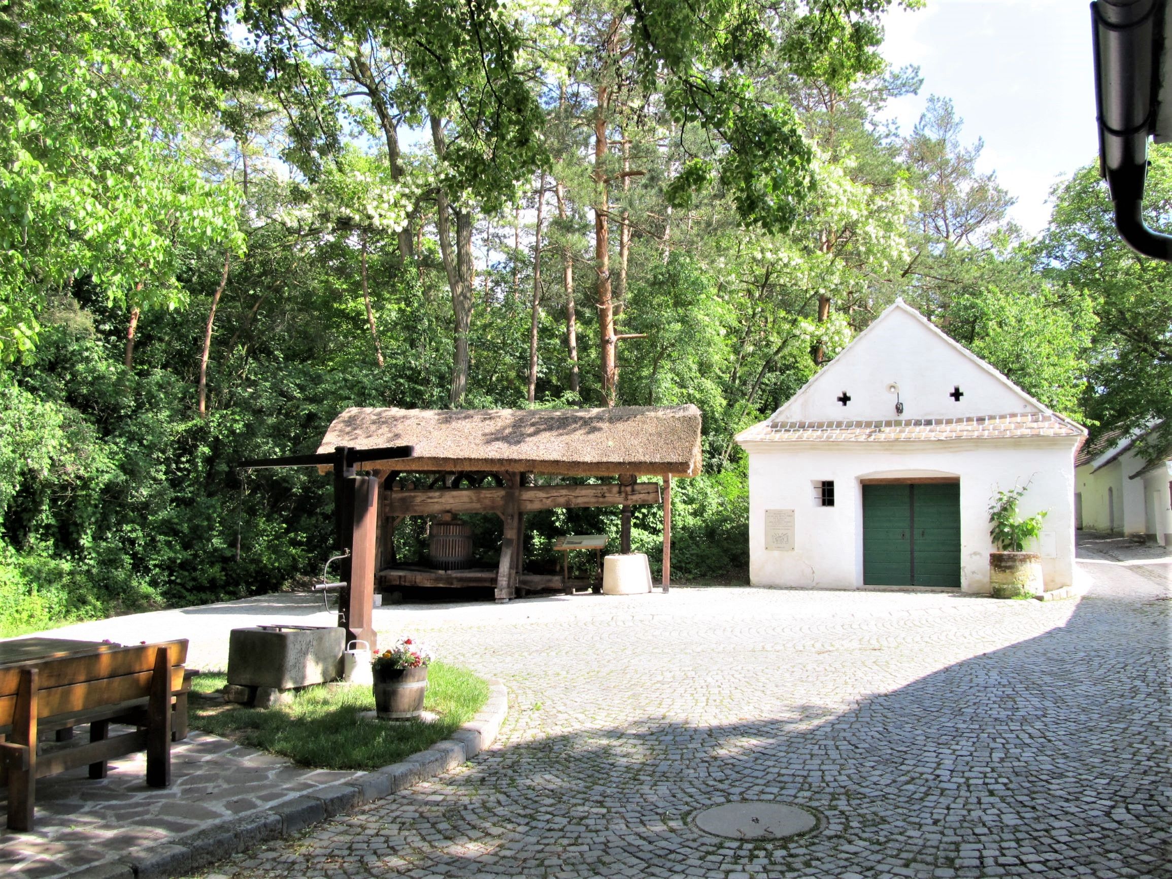 A traditional wine cellar with an old wine press and a small white building in the background, surrounded by trees.