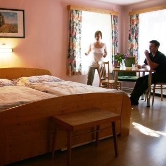 A cozy room with a double bed, table and two people chatting. Sunlight falls through the windows with colorful curtains.