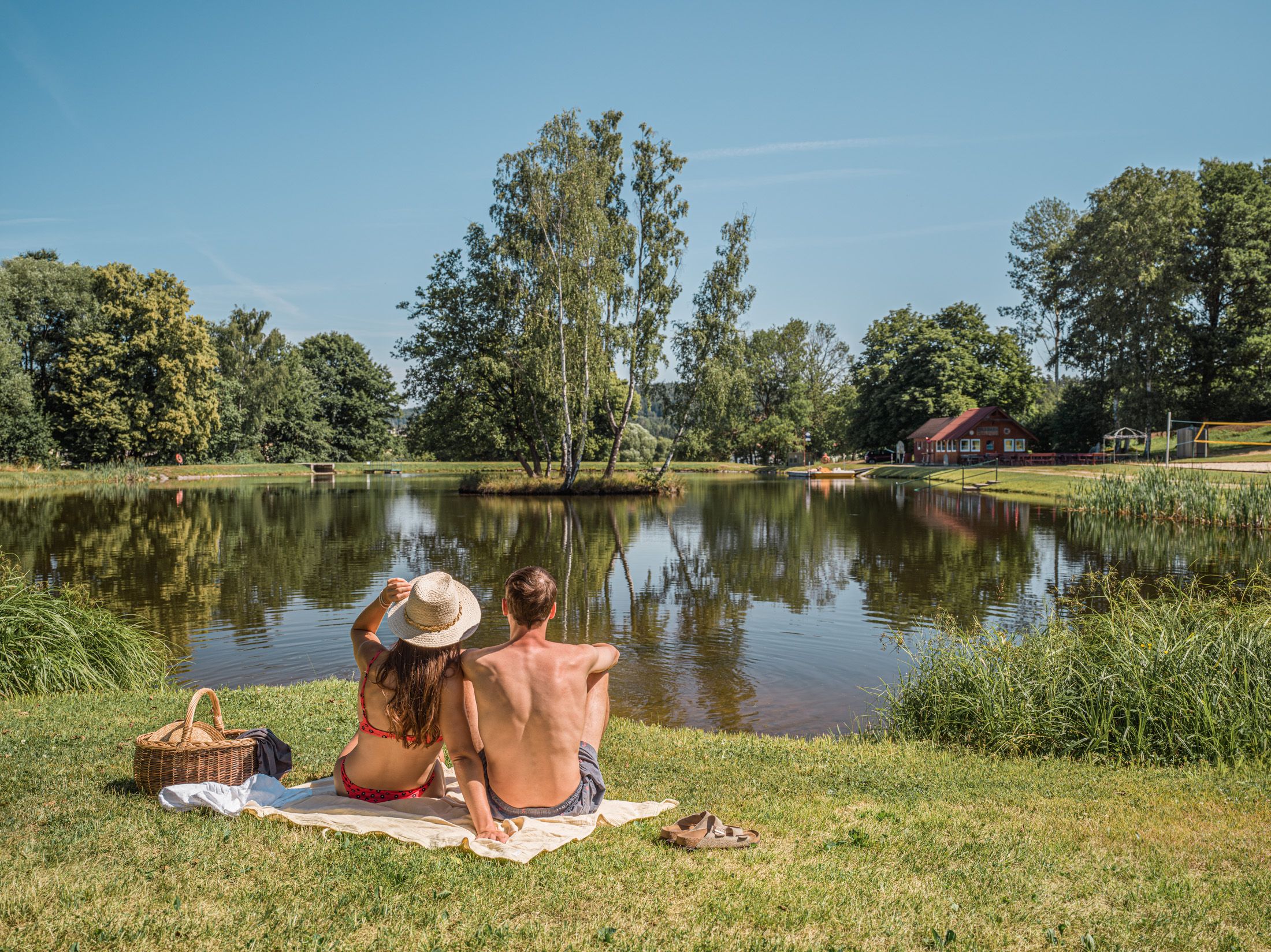 A couple sits on a blanket on the shore of a lake, surrounded by trees and a red building in the background.