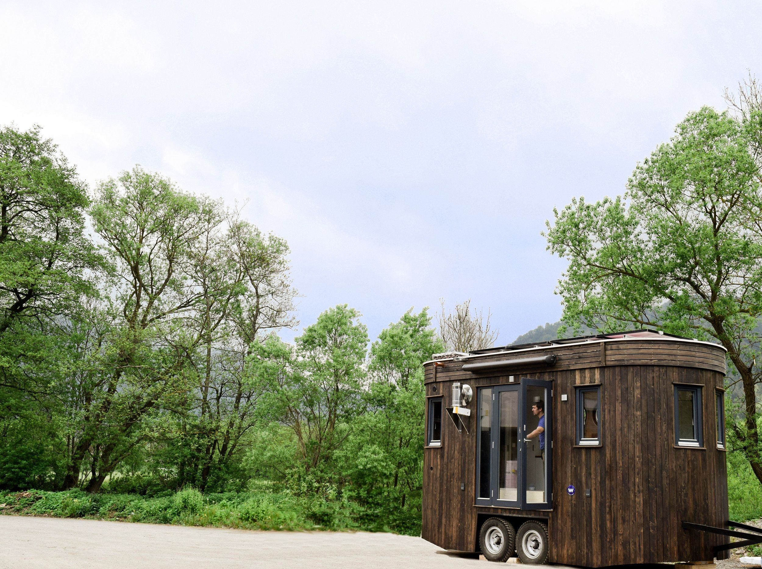 A small, dark caravan with wooden cladding stands in a parking lot in front of green trees and a cloudy sky.