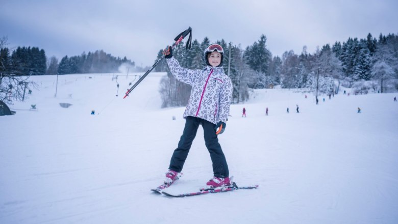 A child in ski equipment stands on a snowy slope and holds ski poles in the air. Trees and other skiers can be seen in the background.