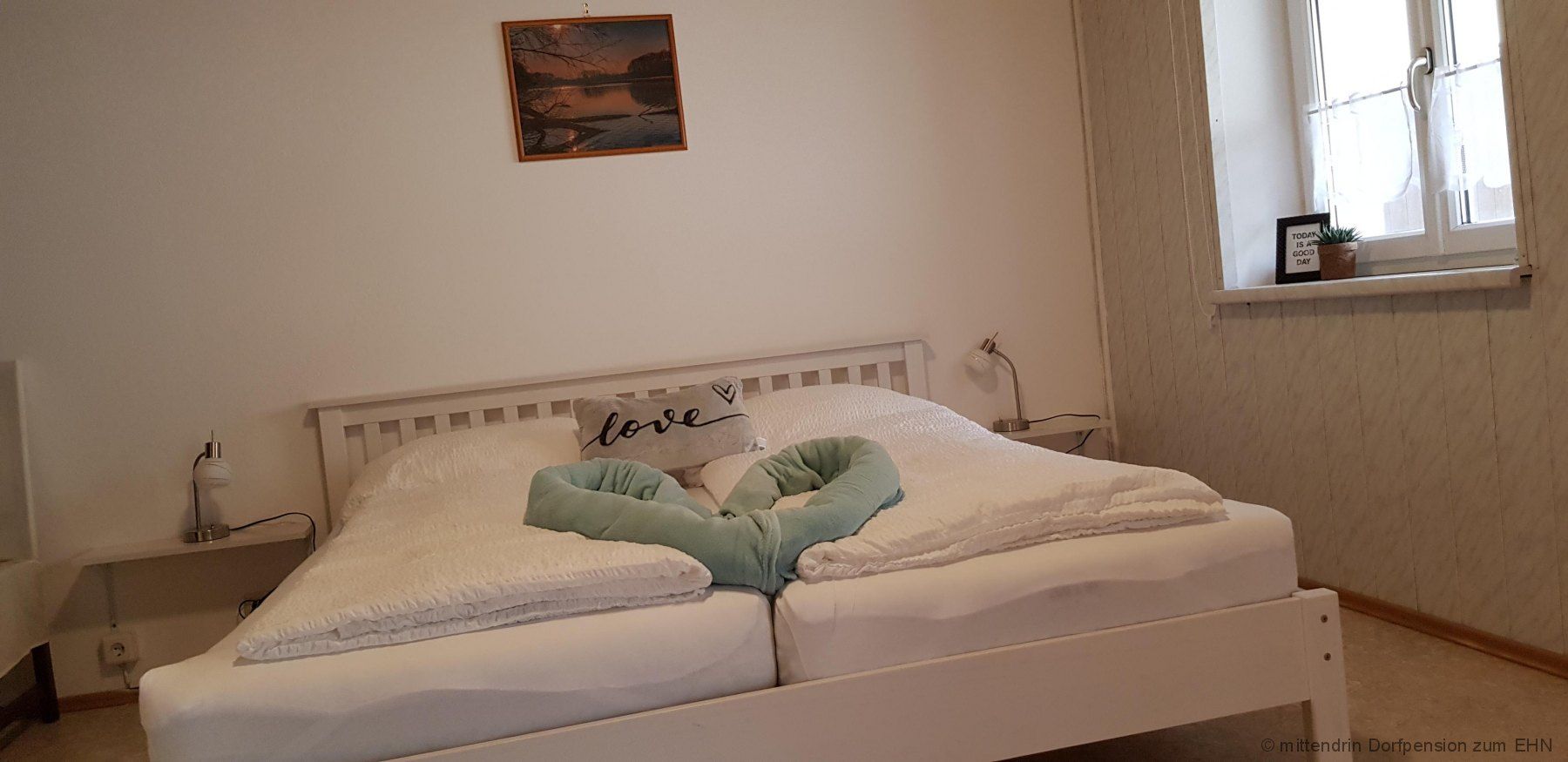 White double bed with a blanket that forms a heart. A red picture hangs on a white wall above the bed. 
