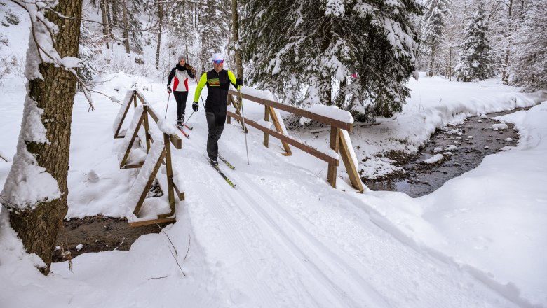 Two people cross a stream in a snowy forest landscape while cross-country skiing