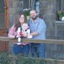 A couple with a baby stands in front of a wooden house.