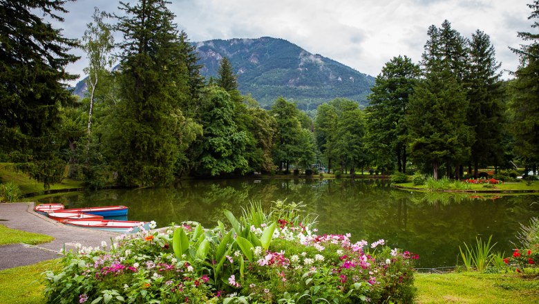 A picturesque park with a pond, surrounded by trees and flowers, with mountains in the background.