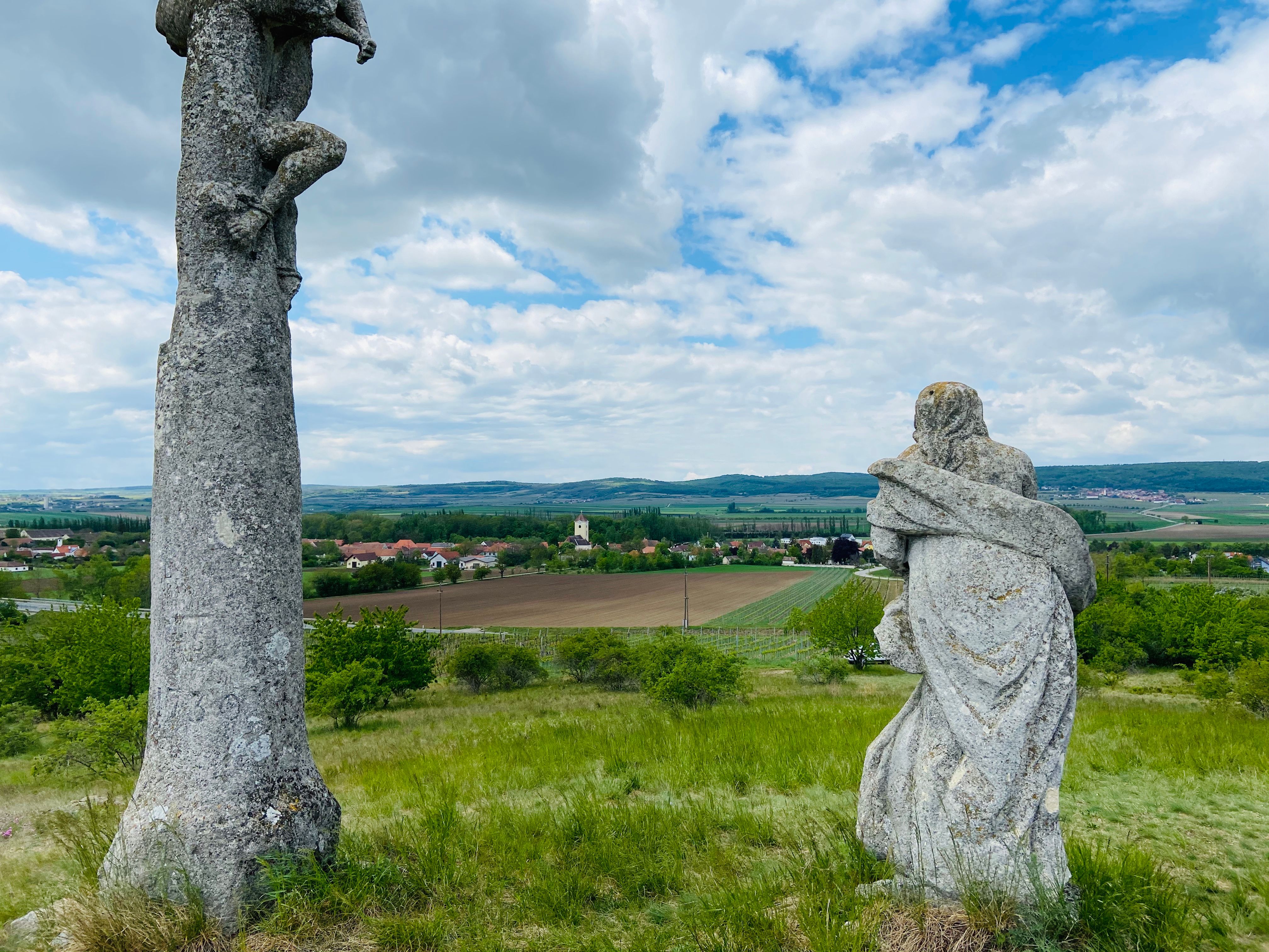 Two stone sculptures on a hill with a view of a rural landscape and a village in the background.
