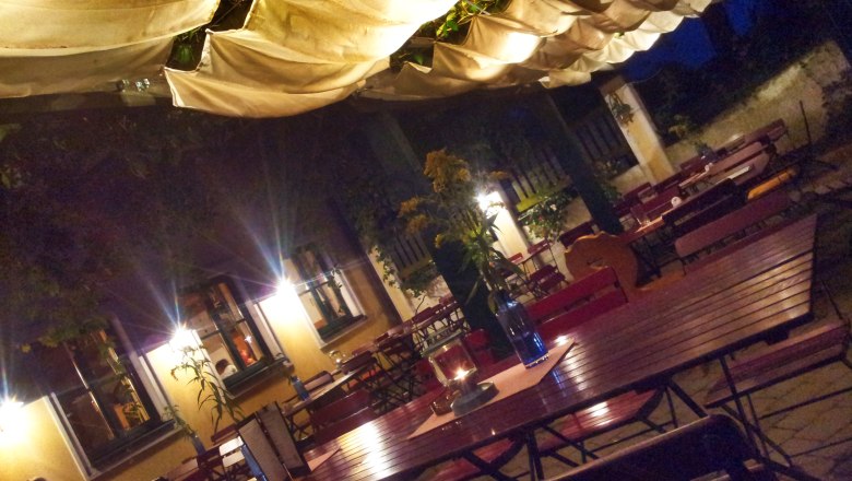 Illuminated terrace with wooden tables and chairs, covered with fabric panels, at night.