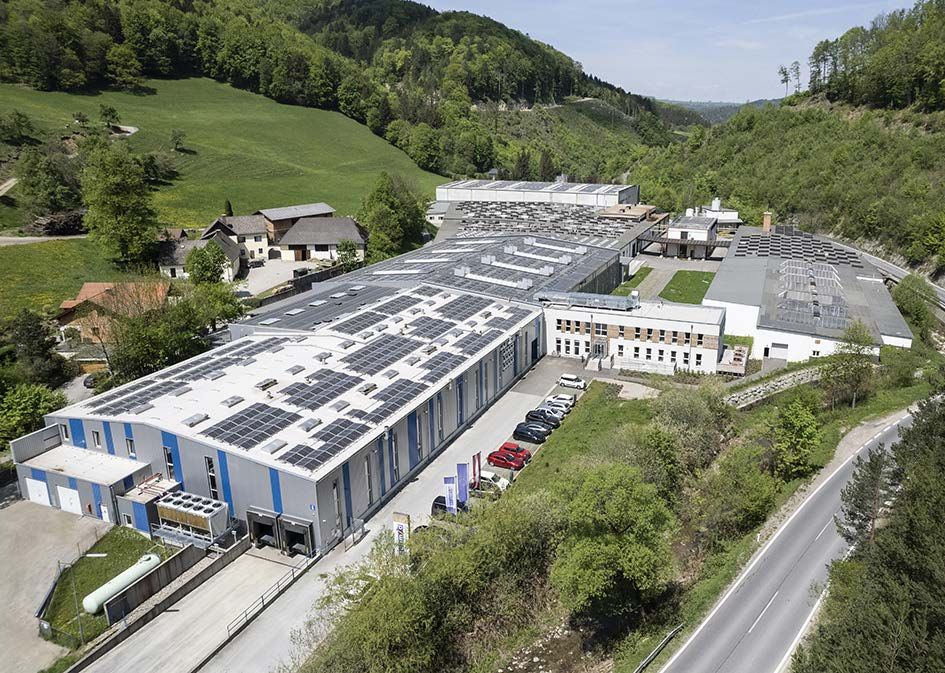 Aerial view of a large factory with solar panels on the roofs, surrounded by a green landscape.