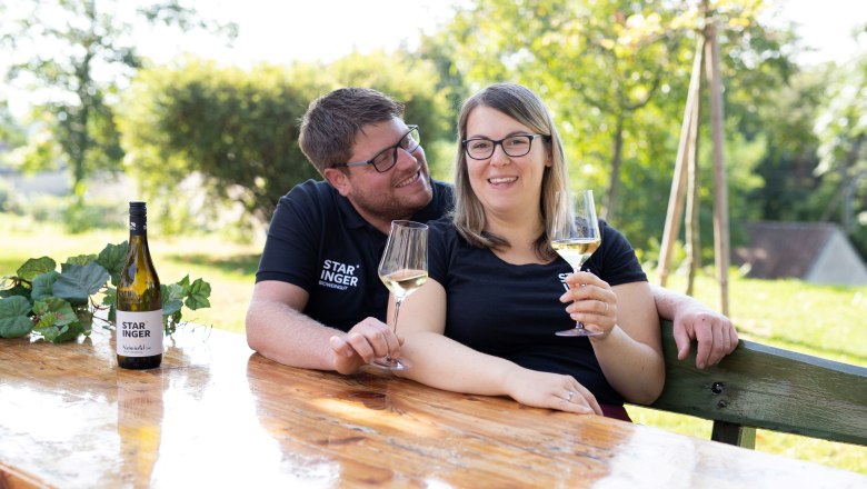 A couple is sitting at a wooden table outside, holding glasses of wine. A bottle of wine with the label 'Staringer' is on the table.