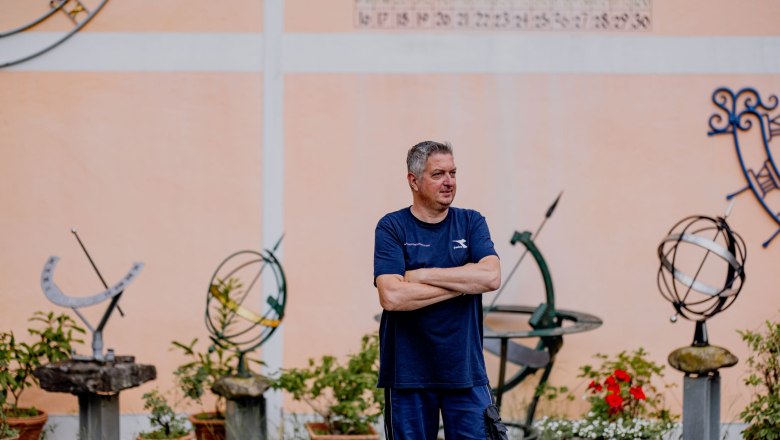 A man stands with his arms folded in front of a wall with sundials and plants.