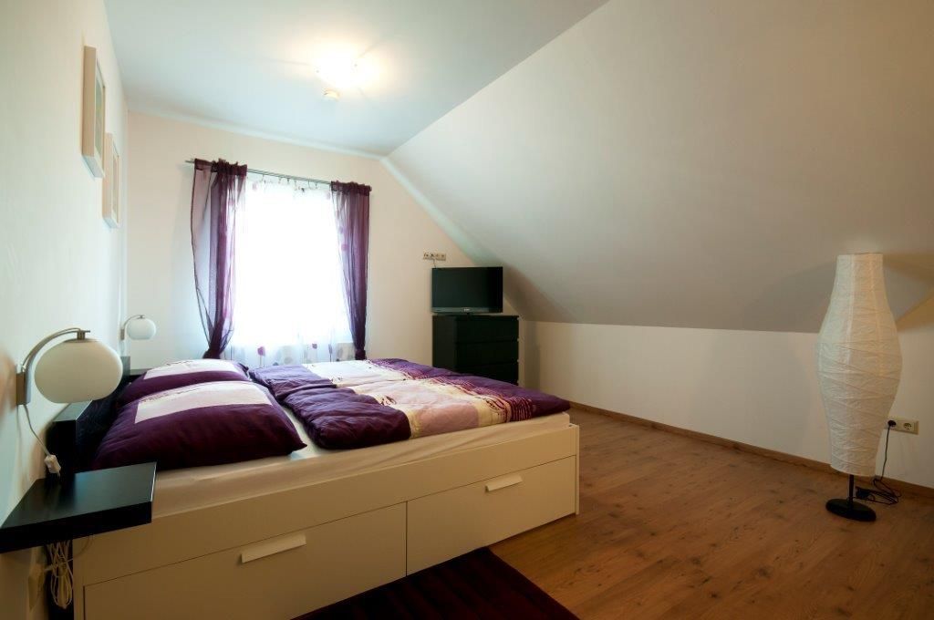 A cozy guest room with a double bed, purple curtains and a TV on a chest of drawers under a sloping roof.