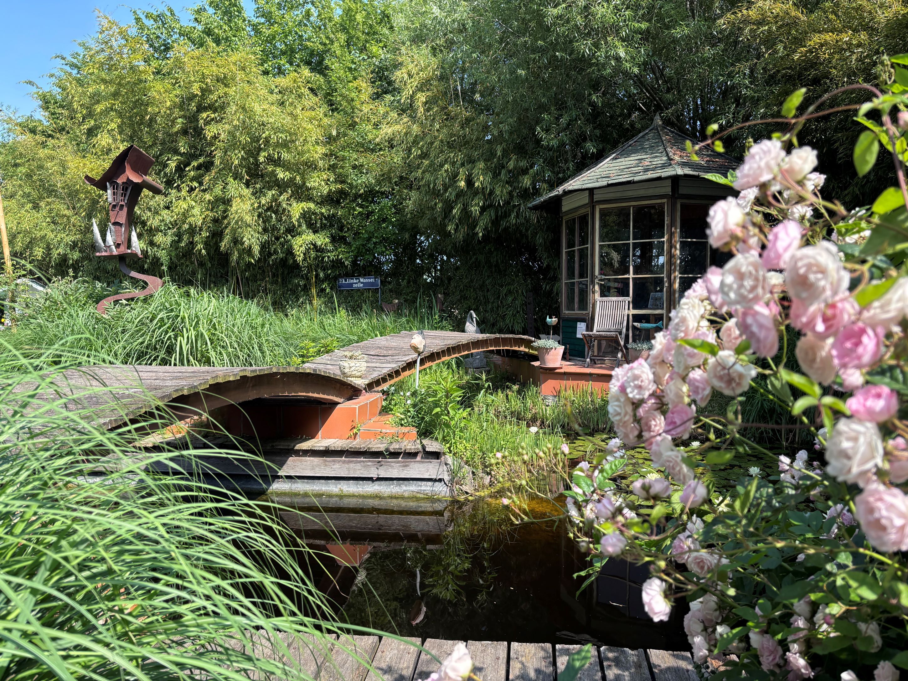 An idyllic garden with a pond, bridge, pavilion and blooming roses in the foreground.