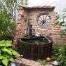 A rustic garden fountain with a brick wall, surrounded by plants and an old wagon wheel as decoration.