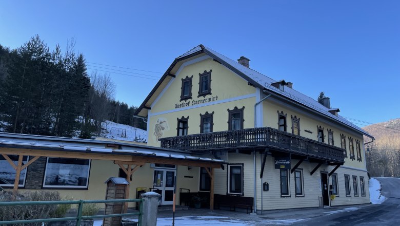 Yellow building of the Karnerwirt inn with wooden balcony, surrounded by snow-covered trees.