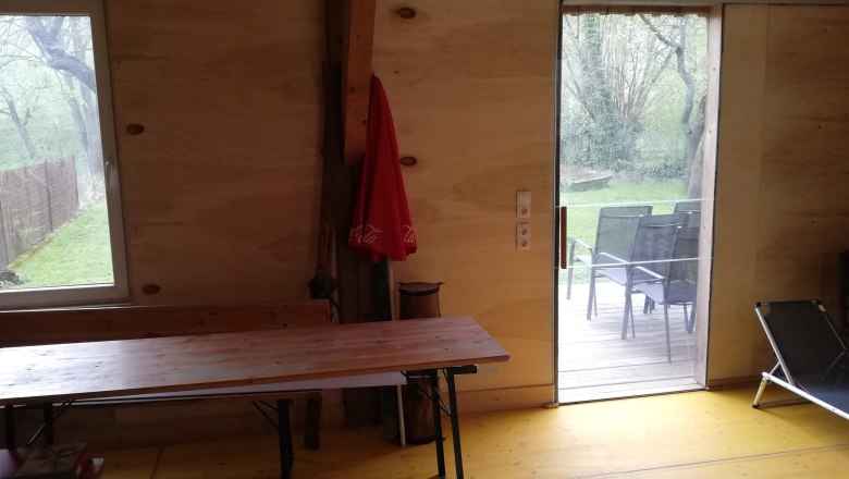 Interior view of a room with wooden walls, table, deck chair and view of the terrace and garden.