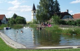 A natural bathing pond with people swimming. A church tower and houses can be seen in the background.