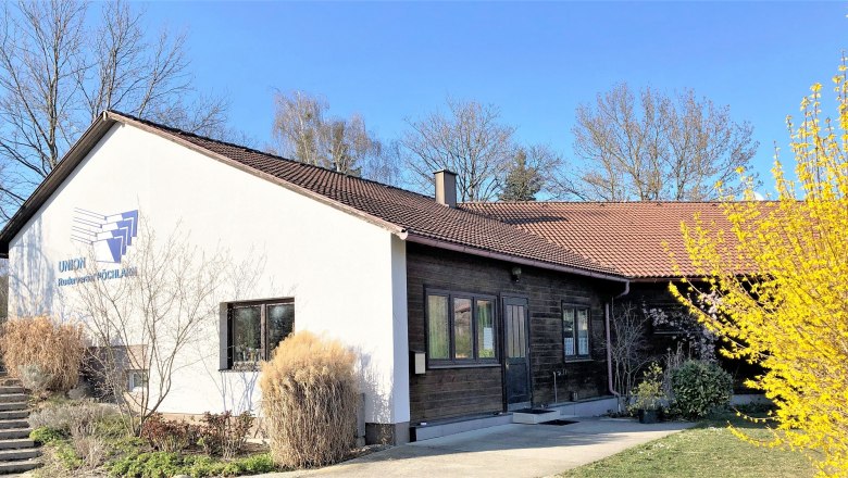 Boathouse of the Union Rowing Club P&ouml;chlarn with white fa&ccedil;ade and brown roof, surrounded by trees and bushes.