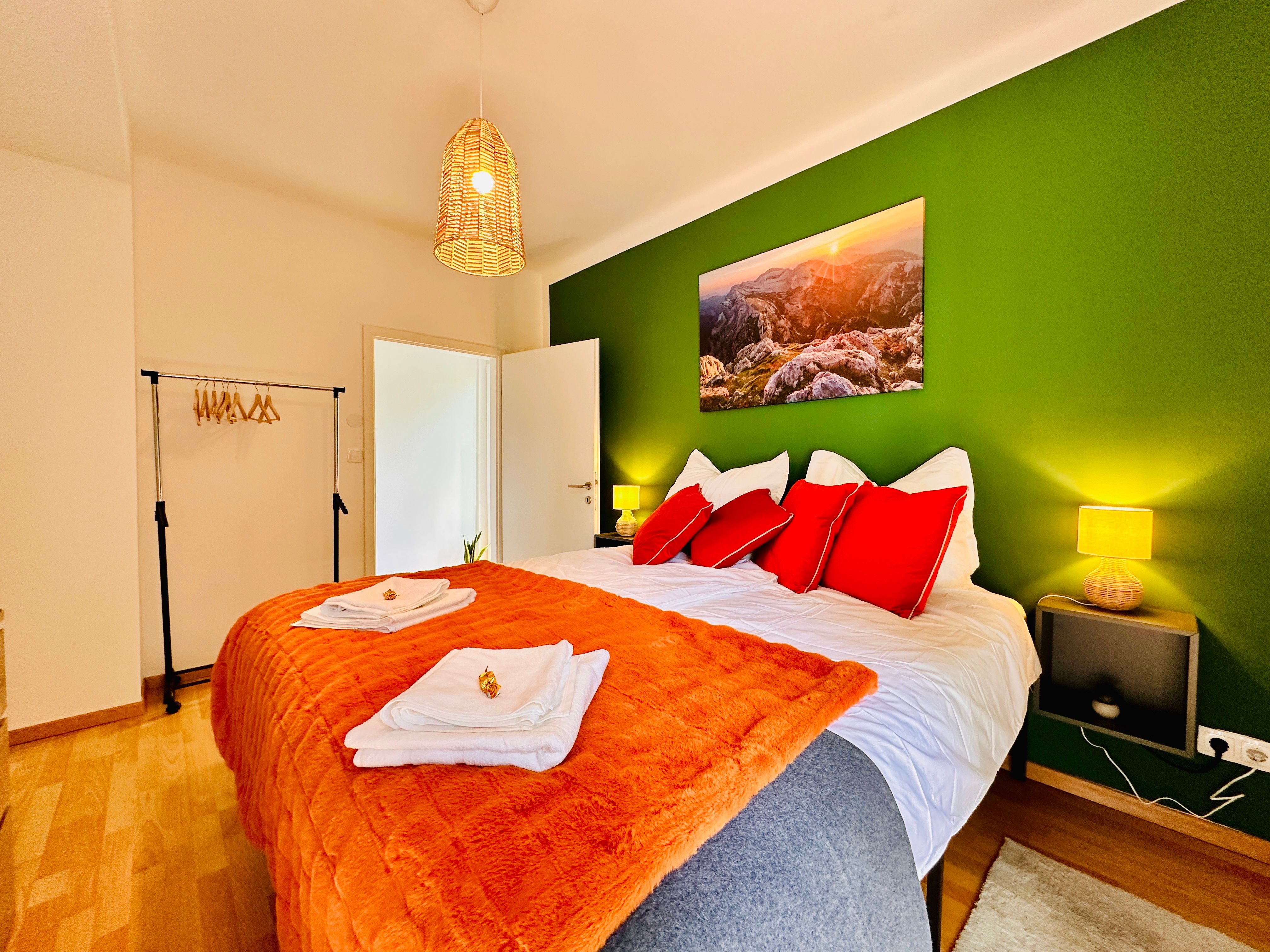 Cozy bedroom with green painted wall, double bed with red pillows, orange blanket and landscape picture. Hanging lamp and clothes rack available.