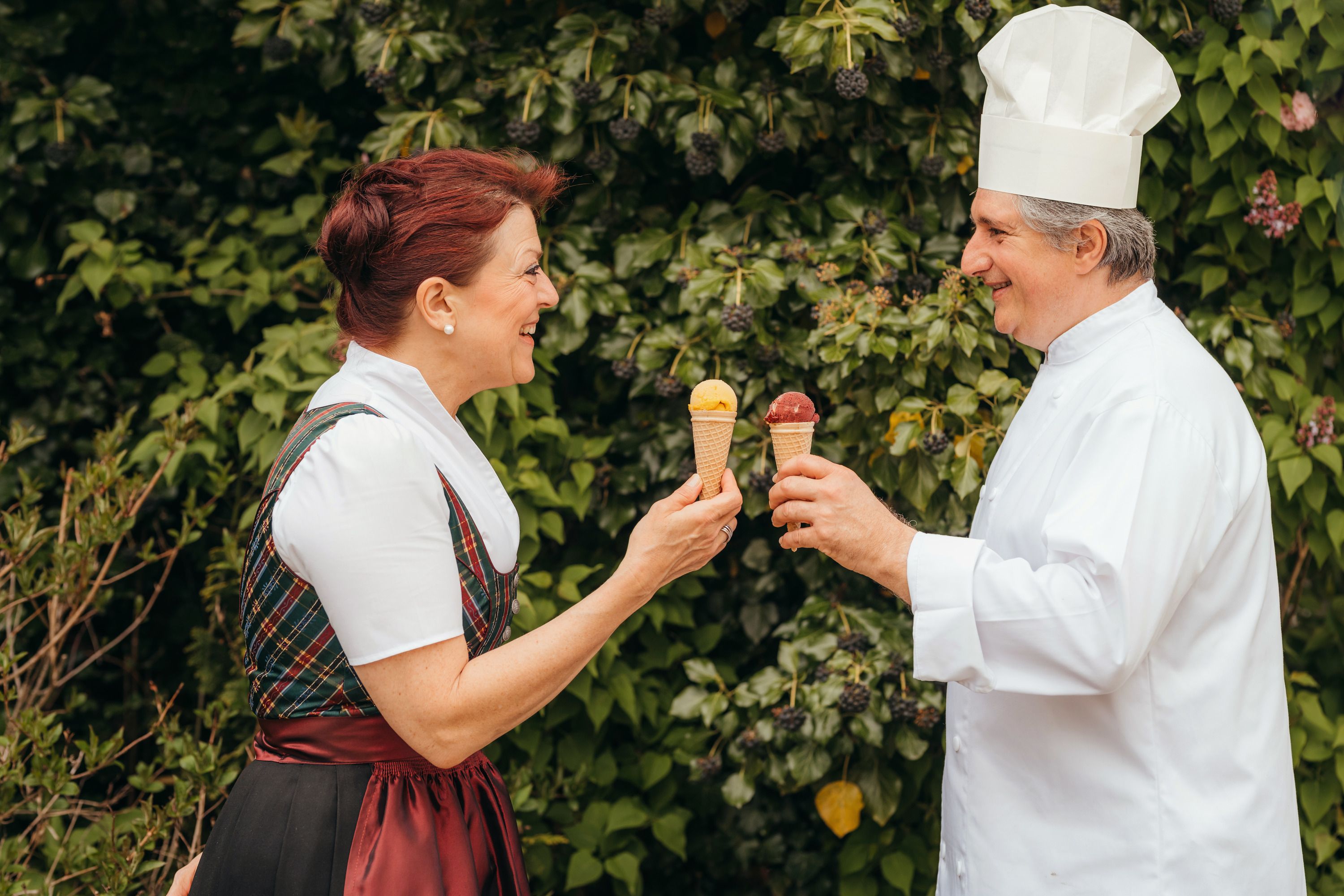 Two people holding ice cream cones and smiling at each other against a background of green leaves.