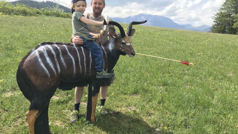 A man holds a child on a 3D target dummy of an antelope-like animal in a meadow with mountains in the background.