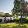 People sit at a table in the garden under a pergola, surrounded by nature and sunshine.