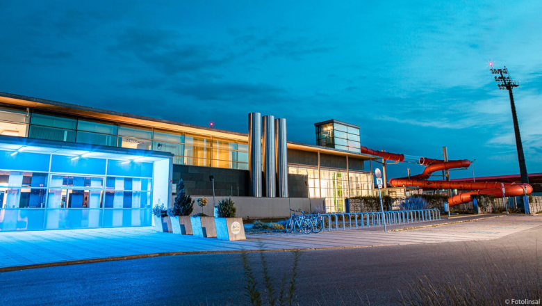 Exterior view of a modern swimming pool at dusk with blue lighting and red water slide.