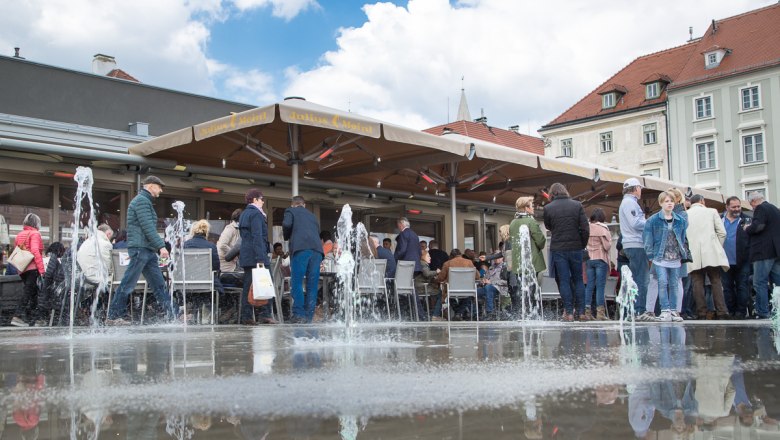 People sit and stand around a fountain in a square with a caf&eacute;.