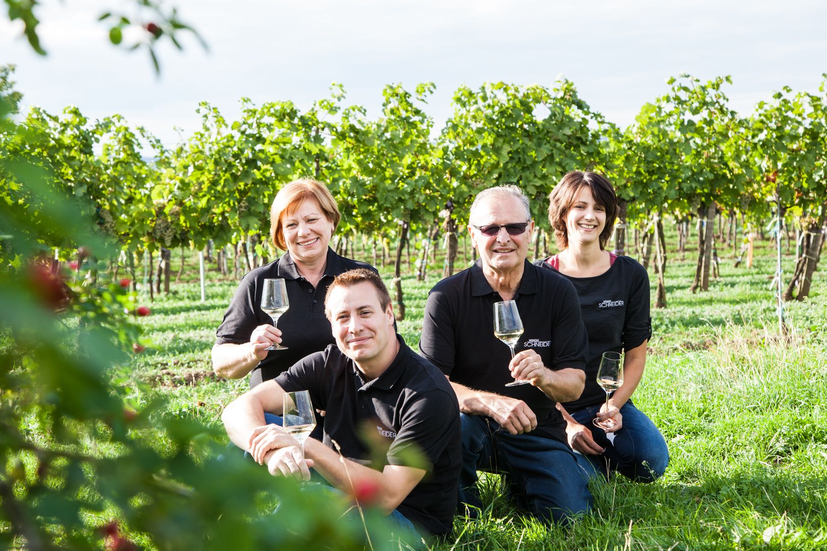 Four people are sitting in a vineyard holding glasses of wine. They are wearing black clothes and smiling at the camera.