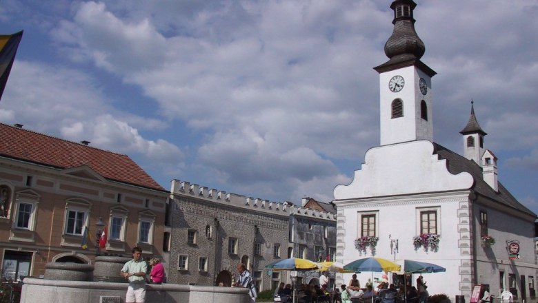 A town square with a white building with a tower and clock, people sitting under parasols, a fountain in the foreground.