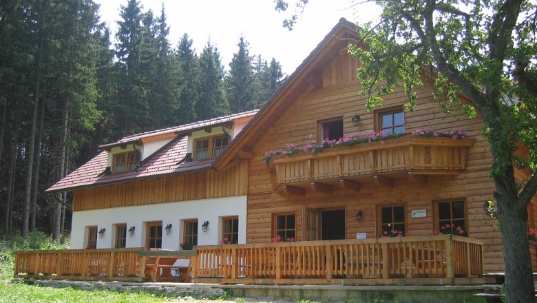 A rustic wooden mountain hut with balcony and terrace, surrounded by trees.
