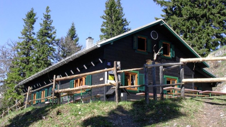 A mountain hut with a wooden fence, surrounded by trees, in sunny weather.