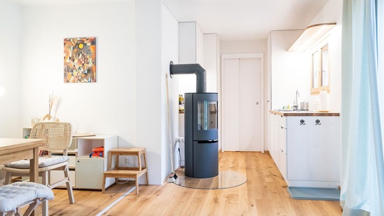 Modern living area with wooden floor, wood-burning stove, dining table and kitchenette.