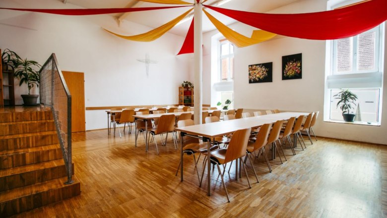 A bright dining room with a wooden floor, tables and chairs. Red and yellow fabric panels hang from the ceiling.