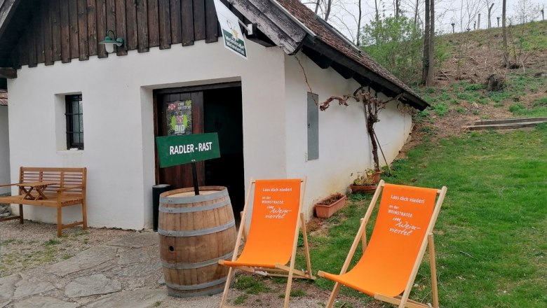 A small white building with a wooden roof, two orange deckchairs and a wooden barrel in front of it, surrounded by trees and grass.