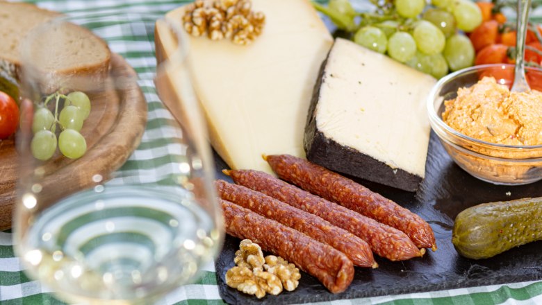 A table laid with cheese, sausage, bread, grapes and a glass of white wine on a green and white checkered tablecloth.