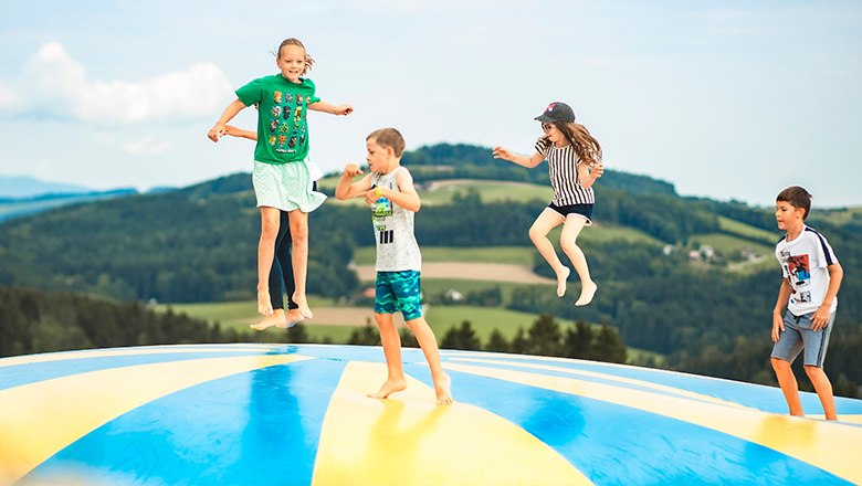 Four children jump on a colorful bouncy cushion in front of a hilly landscape with lots of trees.