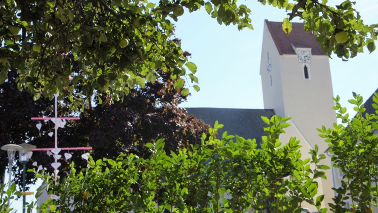 A church tower behind green trees and bushes, with a decorative element in the foreground.