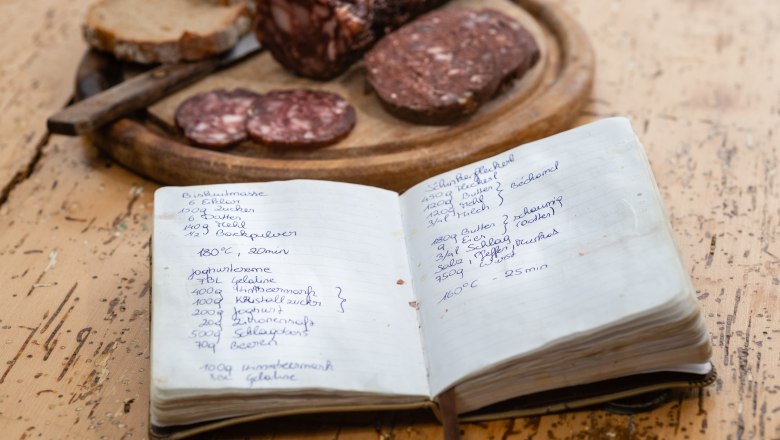 A handwritten recipe book lies on a table next to a wooden platter with venison sausage and bread.