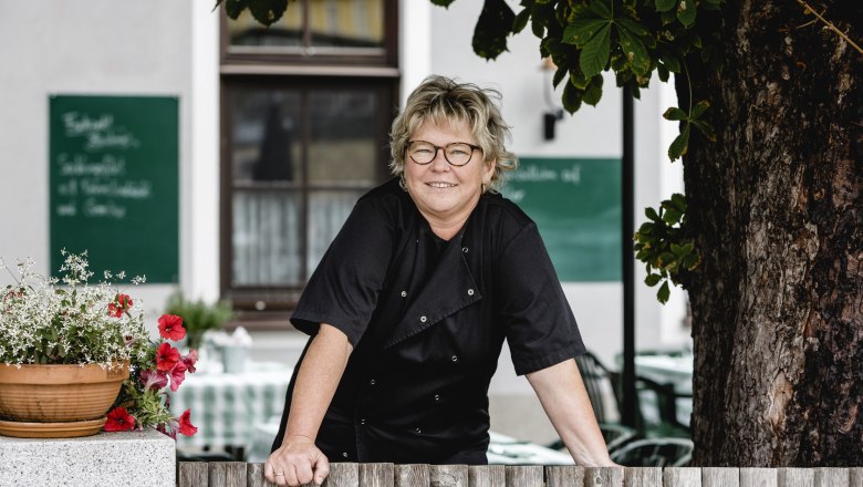 A woman in black clothing leans against a wooden fence, with flowers and a tree in the background.