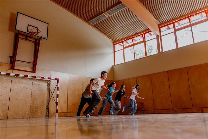 Five teenagers running in a gym with a basketball hoop and handball goal.