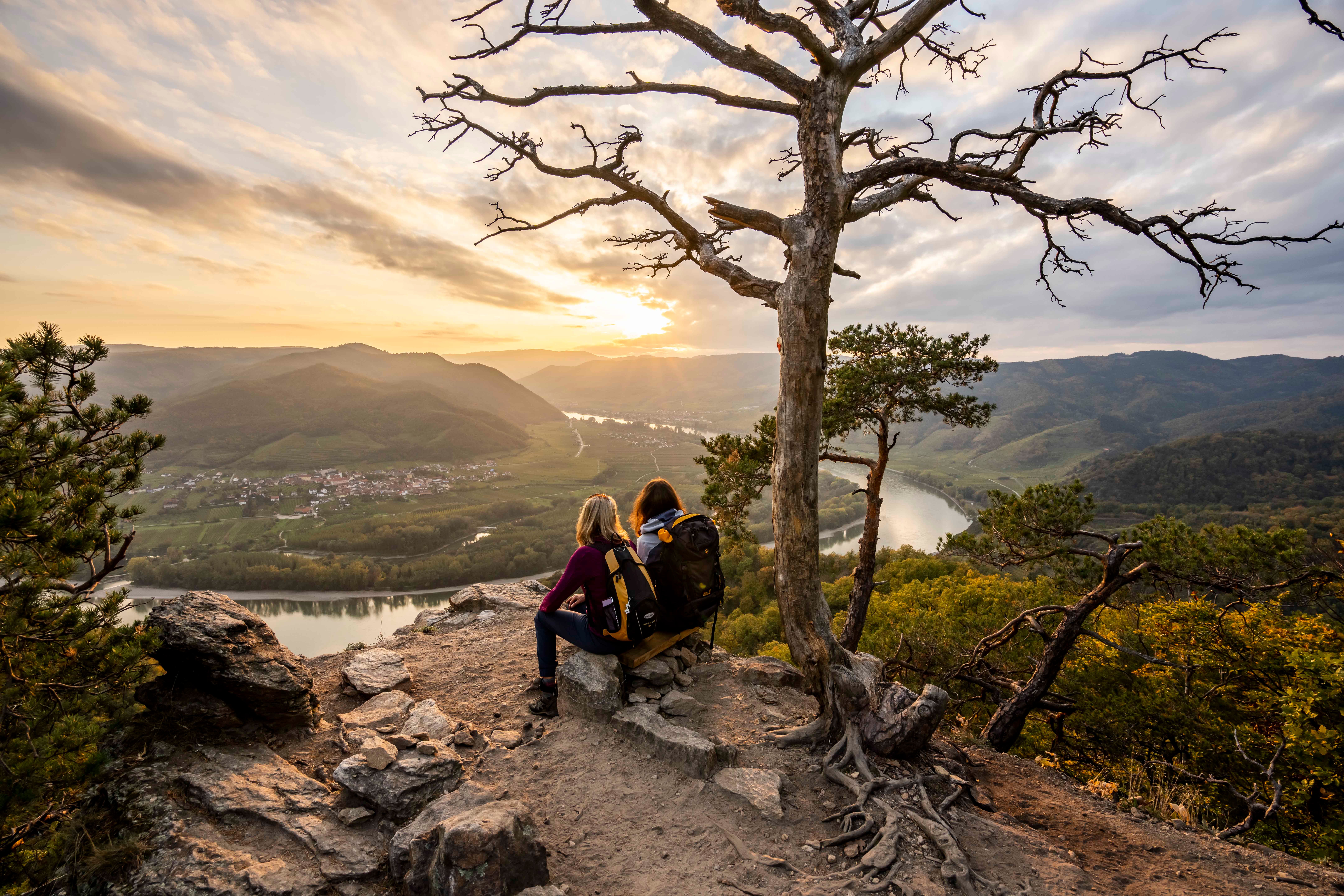 Two people sit on a rock and look out over a river landscape at sunset.