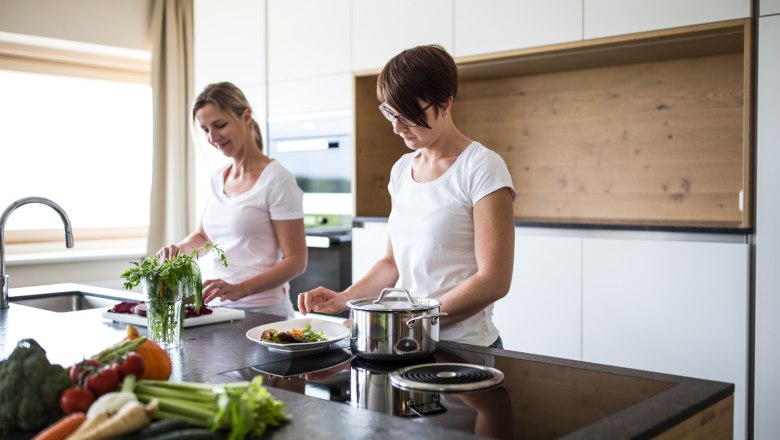 Two women cooking in a modern kitchen with fresh vegetables on the worktop.