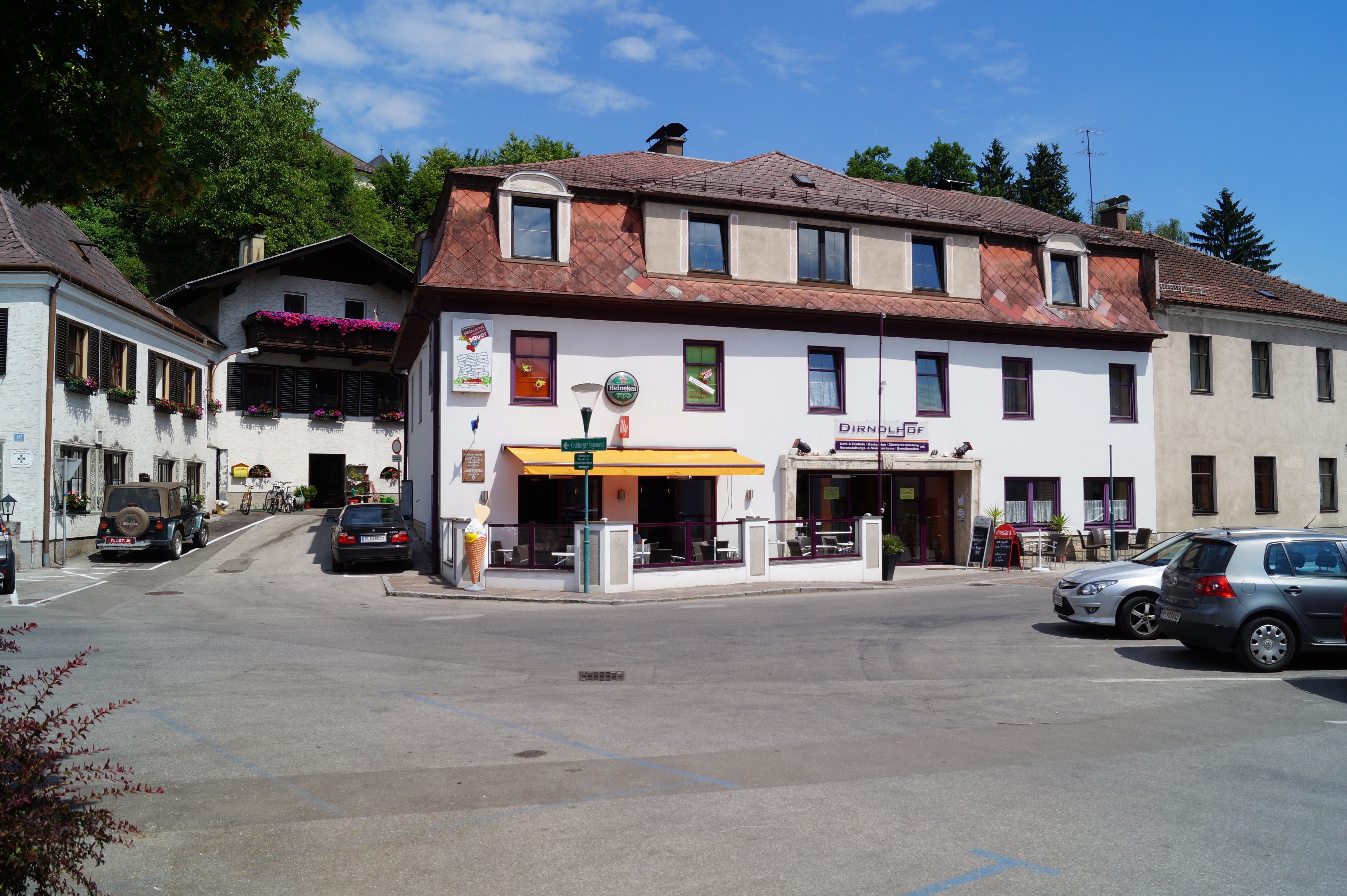 A building with the sign 'Dirndlhof' and a terrace with a parasol. Cars are parked in the parking lot in front of it.