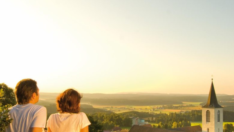 Two people sit on a rock and look out over a church and a vast landscape at sunset.