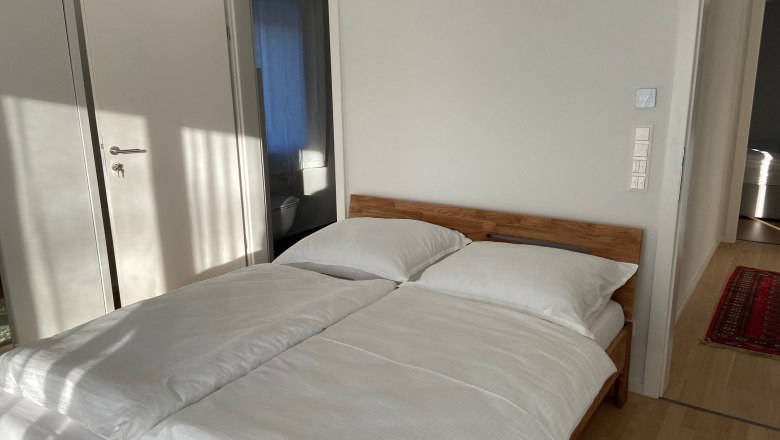 One bedroom with a double bed, white comforter covers and wooden floor. An open door leads to a bathroom.