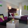 Modernly furnished hotel room with double bed, sofa and decoration in shades of purple and green.