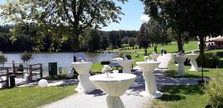 Bar tables with white cloths on the shore of a lake, surrounded by trees.