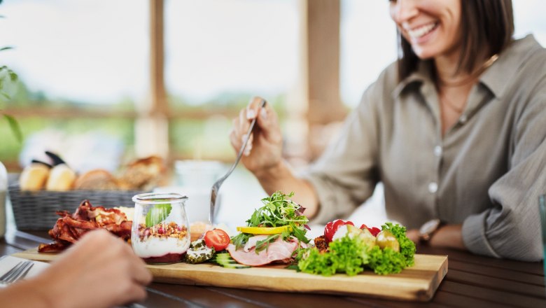A woman eats breakfast with fresh ingredients on a wooden board on a terrace.