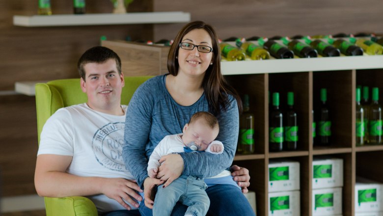 A couple with a baby sits on a green armchair in a winery.