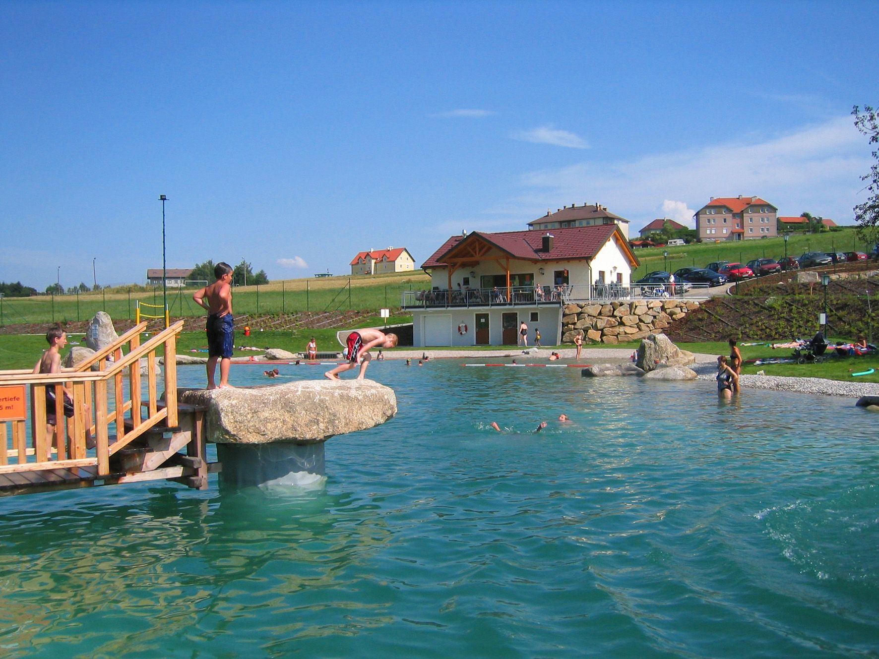 Children playing at a swimming pond with a jetty and rocks, a building and cars in the background.